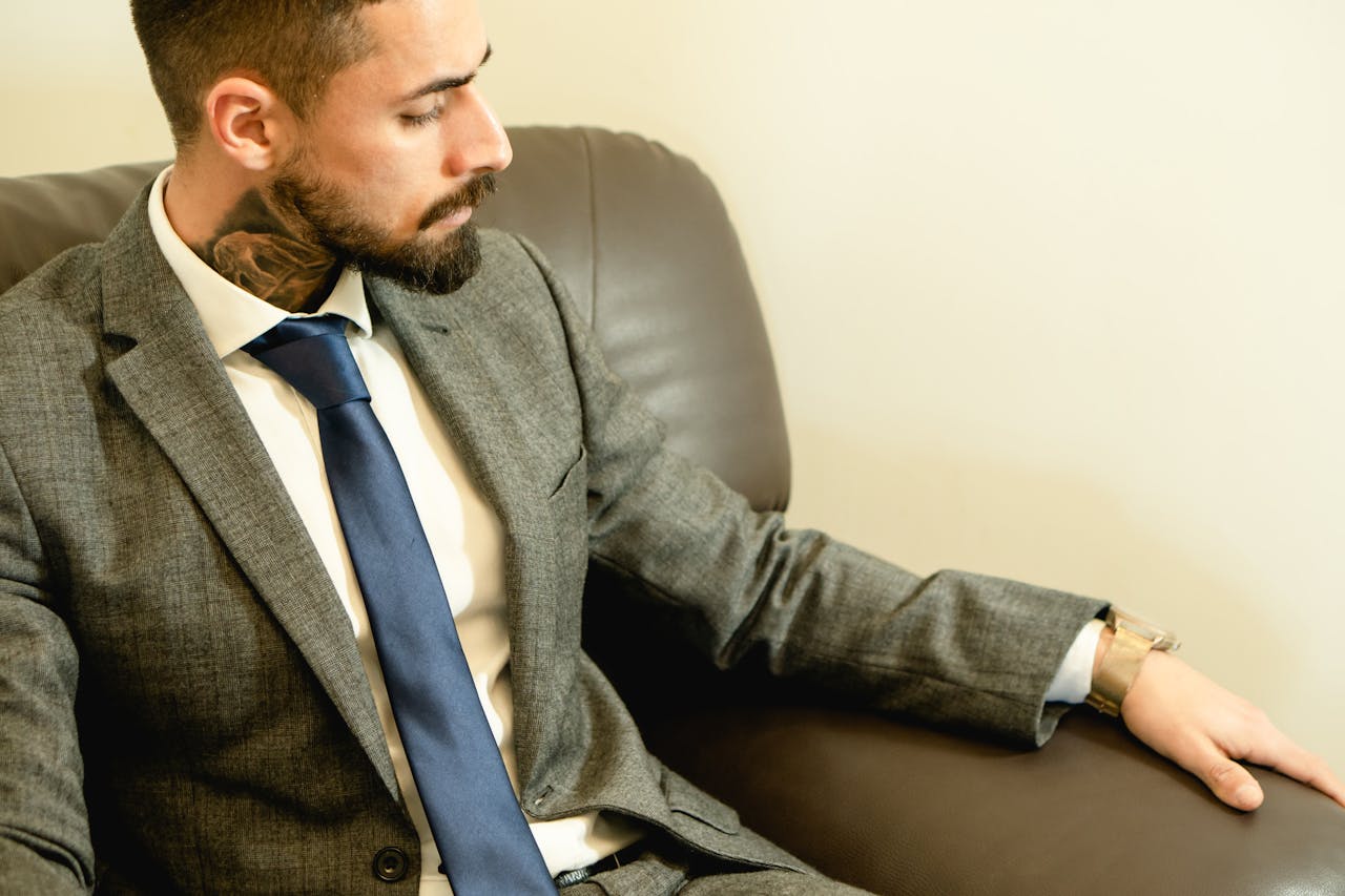 Caucasian man with beard and neck tattoo in a grey suit seated indoors, exuding elegance.